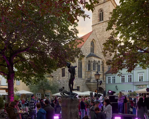 Kirche und Brunnenfigur über Gästen auf einem violett beleuchteten Platz in der Abenddämmerung.