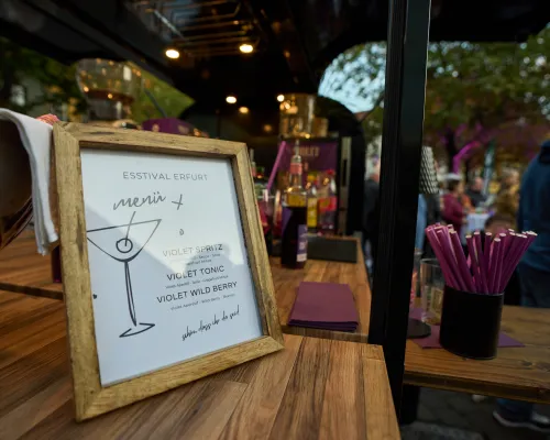 Eine gerahmte Getränkekarte steht neben violetten Trinkhalmen auf einer hölzernen Bar.