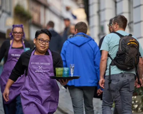 Ein Teilnehmer in violetter Schürze balanciert ein Tablett mit blauen und grünen Gläsern sowie einem Sektglas.