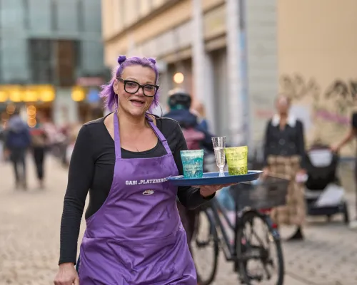 Eine Frau mit violettem Haar und Brille trägt in einer Schürze ein Tablett mit Getränken.