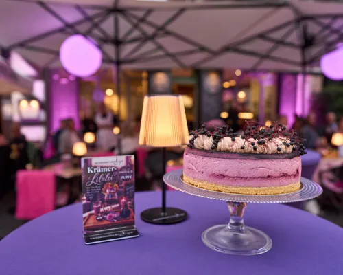 Eine Beerentorte steht neben einer warm leuchtenden Tischlampe auf einer violetten Tischdecke.