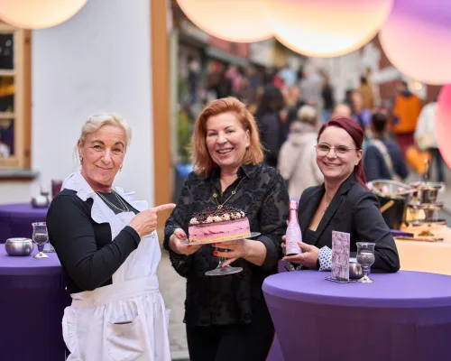 Drei lächelnde Frauen präsentieren eine Torte und eine rosafarbene Getränkeflasche.