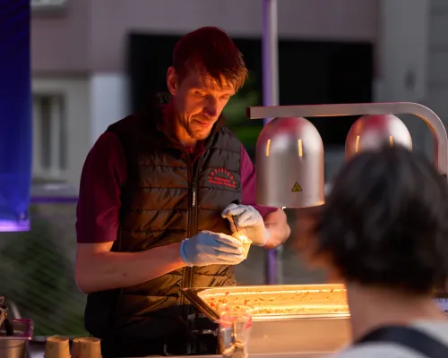 Ein Mann portioniert Speisen unter zwei Wärmelampen; vor der Theke steht ein Gast.