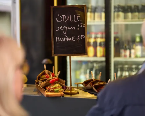 Eine Kreidetafel mit veganen und rustikalen Stullen steht hinter belegten Broten an einer Verkaufstheke.