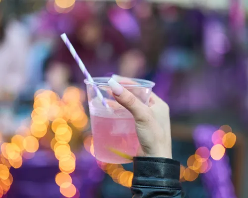 Eine Hand hält ein Glas mit rosafarbenem Getränk, Zitrusscheibe und gestreiftem Trinkhalm vor unscharfen Lichtern.