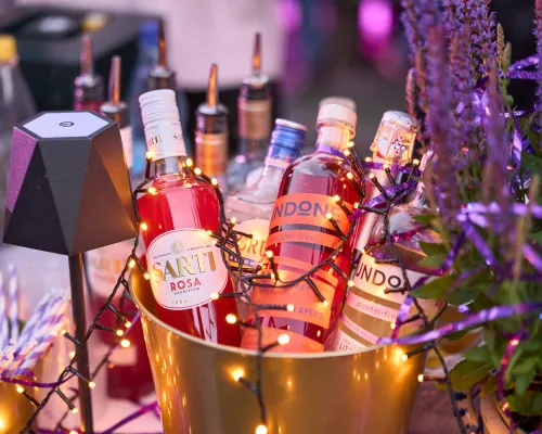 Beleuchtete Aperitifflaschen stehen in einem goldfarbenen Kühler neben Blumen.
