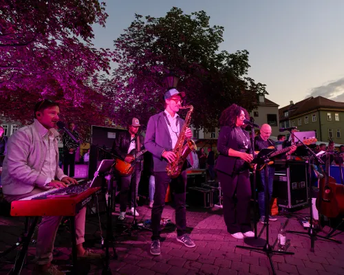 Eine Band mit Keyboard, Saxophon, Gesang und Bass spielt vor violett beleuchtetem Hintergrund in der Dämmerung.