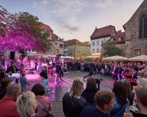 Tanzende treten auf einem violett beleuchteten Platz auf, umringt von zuschauenden Gästen.