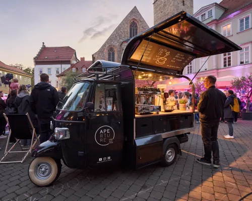 Schräger Blick auf einen schwarzen Kaffeewagen mit geöffnetem Verkaufstresen und Gästen ringsum.