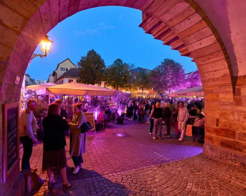 Durch einen Steinbogen fällt der Blick auf den violett beleuchteten Festbereich unter dunkelblauem Abendhimmel.