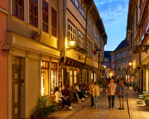 Menschen flanieren durch eine abendliche Altstadtgasse neben besetzten Café-Tischen.