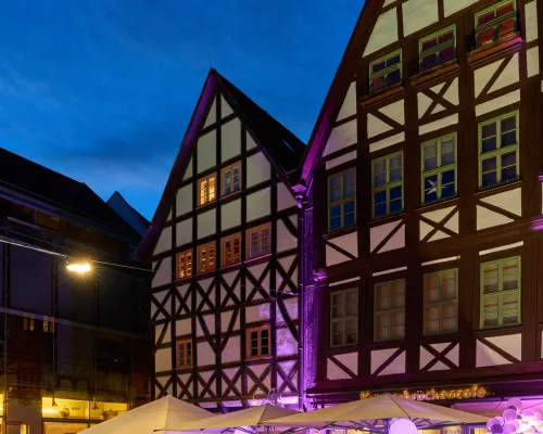 Fachwerkhäuser und eine mit violetten Dekorationen geschmückte Café-Terrasse unter blauem Abendhimmel, im Hochformat.