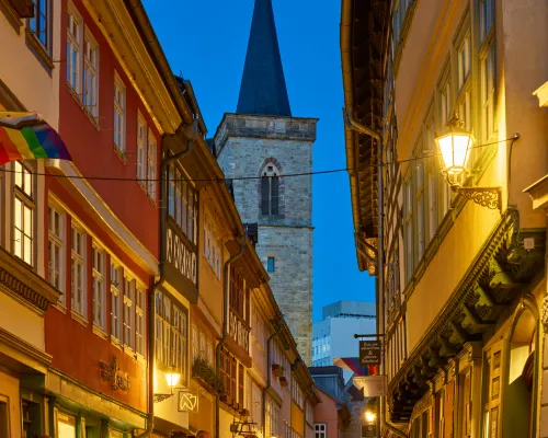 Menschen gehen durch eine beleuchtete Altstadtgasse; dahinter erhebt sich ein Kirchturm.