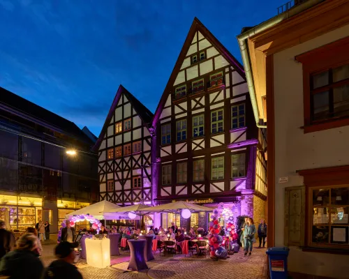 Eine Café-Terrasse mit violetten Luftballons vor Fachwerkhäusern zur blauen Stunde.