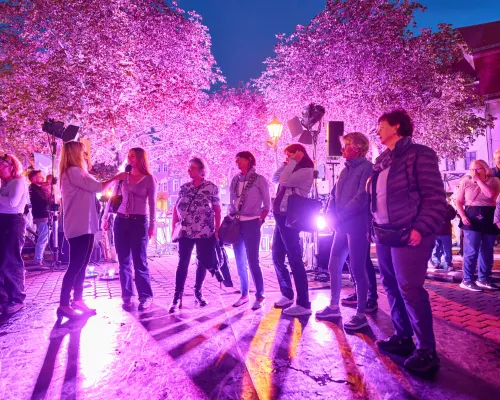 Mehrere Frauen stehen auf einem violett beleuchteten Platz; links hält eine Frau einer anderen ein Mikrofon hin.
