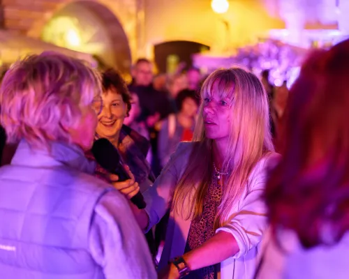 Eine blonde Frau in hellem Blazer hält einer Besucherin ein Mikrofon hin; weitere Gäste stehen daneben.
