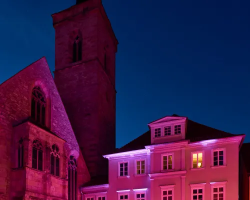 Eine violett beleuchtete Kirche und benachbarte Häuser über einer großen Menschenmenge unter dunkelblauem Himmel.