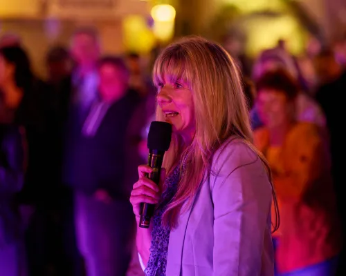Eine blonde Frau in hellem Blazer spricht mit einem Mikrofon vor dem unscharfen Publikum im violetten Festlicht.