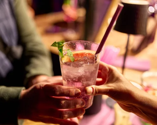 Zwei Hände überreichen ein Glas mit Eis, Minze, einer Grapefruitscheibe und violettem Trinkhalm.