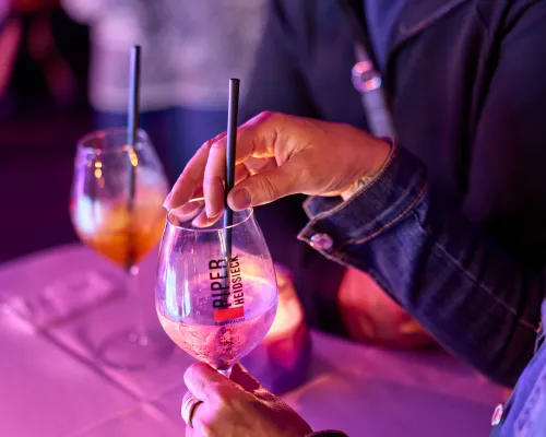 Eine Hand bewegt einen dunklen Trinkhalm in einem Stielglas mit hellem Getränk auf einem violett gedeckten Tisch.