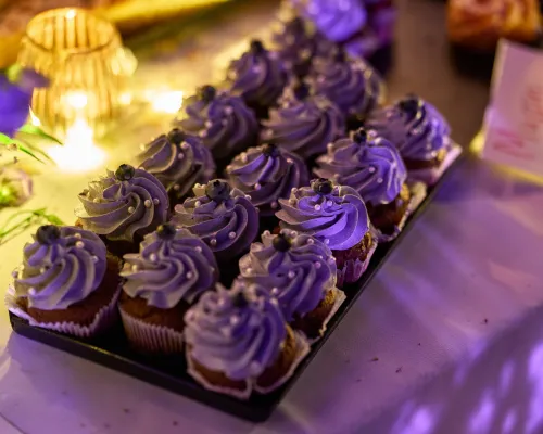 Cupcakes mit violetten Cremehauben, Beeren und kleinen Zuckerperlen auf einem Tablett neben warmem Kerzenlicht.