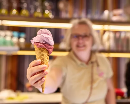 Eine Hand hält eine Waffeltüte mit rosa und violetten Eiskugeln vor einer unscharf sichtbaren Frau an der Theke.