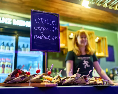 Eine Kreidetafel mit veganen und rustikalen Stullen steht über belegten Broten; dahinter lächelt eine Verkäuferin.