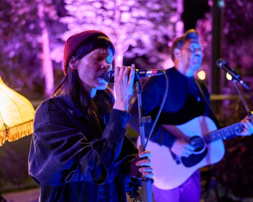 Eine Sängerin mit roter Strickmütze singt am Mikrofon; hinter ihr spielt ein Gitarrist im blauen und violetten Licht.