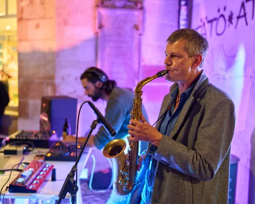 Ein Saxophonist in grauem Jackett spielt vor einer violett beleuchteten Steinwand; hinter ihm bedient ein DJ die Musiktechnik.