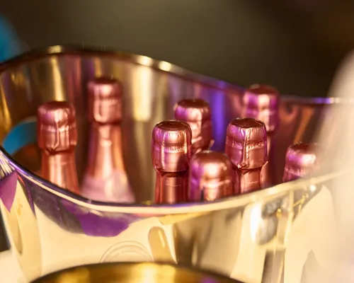 Ein metallener Kühler mit rosafarben umhüllten Flaschenhälsen und warmen Lichtreflexen, im Hochformat aufgenommen.