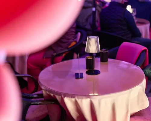 Eine kleine Tischlampe steht mit schwarzen Bechern auf einem gedeckten Tisch zwischen Gästen und pinkfarbenen Decken.