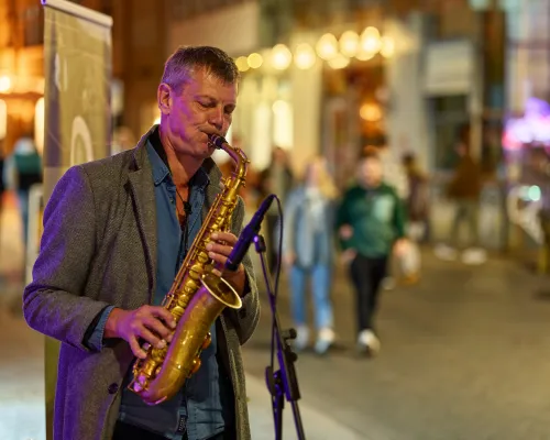 Ein Saxophonist in grauem Jackett und blauem Hemd spielt vor einer unscharfen Altstadtgasse mit warmen Lichtern.